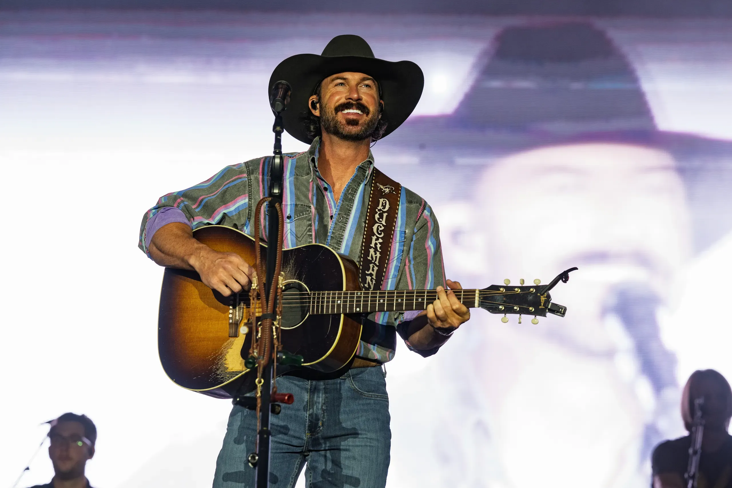 A performer playing acoustic guitar on stage in a hat and patterned shirt.