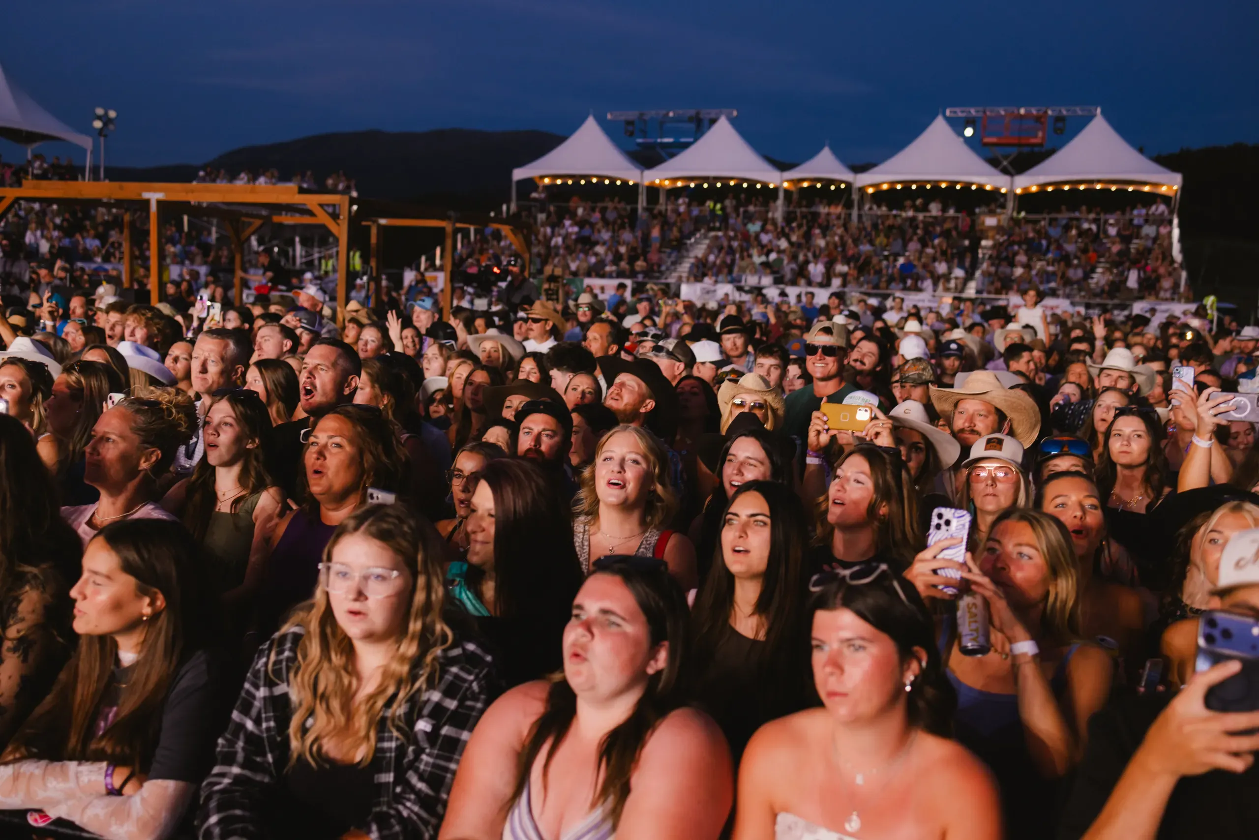 A concert crowd watching the stage at night beneath white event tents.
